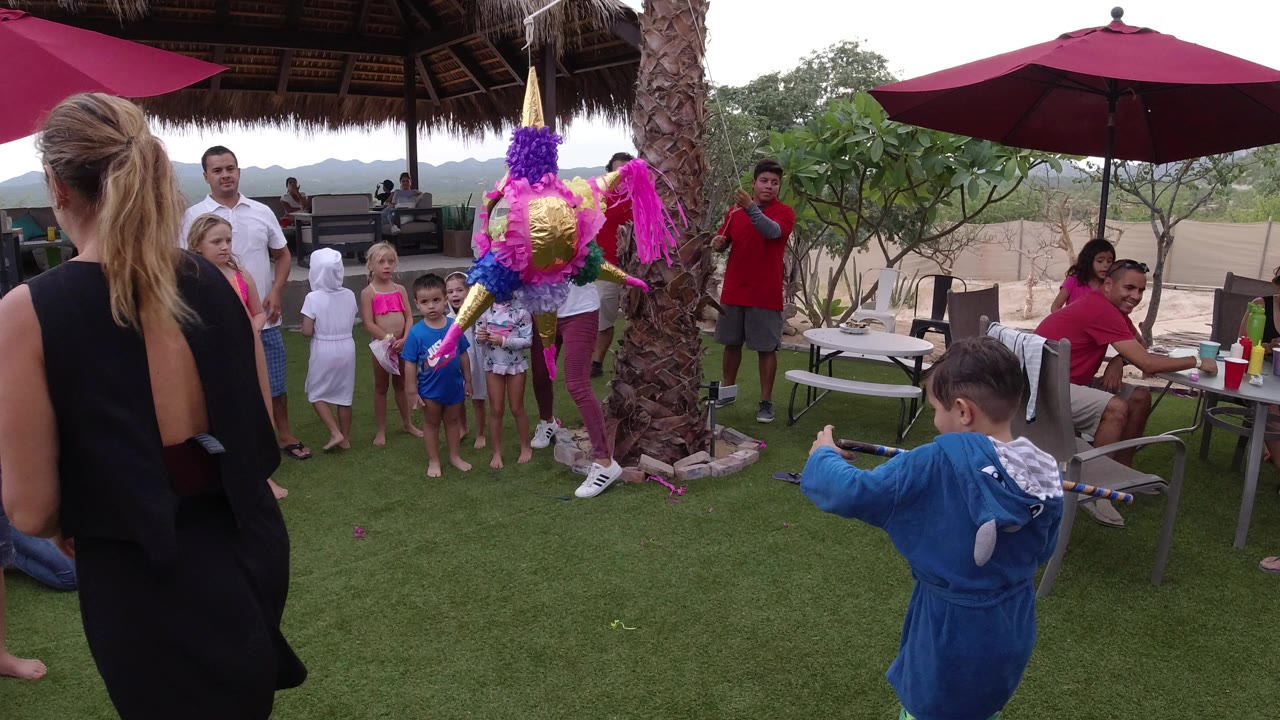 In Los Cabos, Mexico, children and adults gather under a thatched patio roof as a bright piñata hangs beside a palm trunk and a child in a blue robe holds a stick in the foreground.
