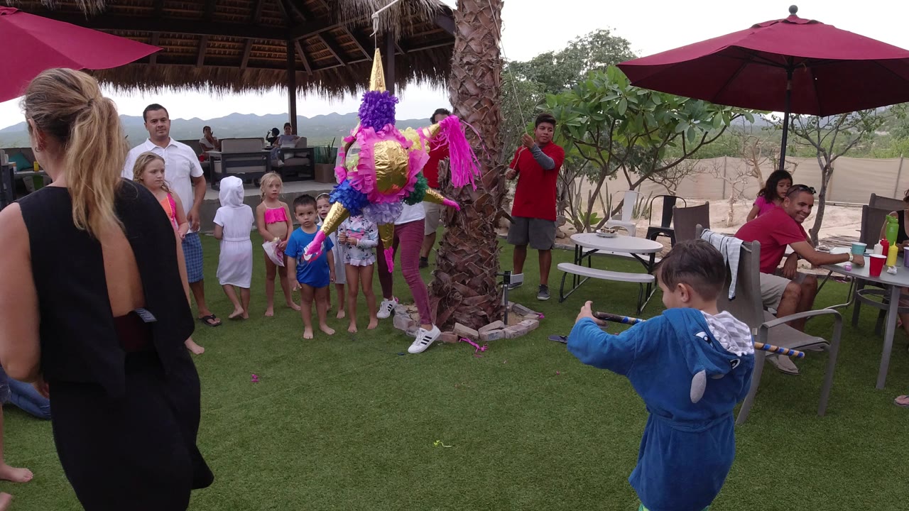 Children line up near the hanging piñata in Los Cabos, Mexico, while adults stand and sit around the shaded patio and a child in blue prepares with a stick.
