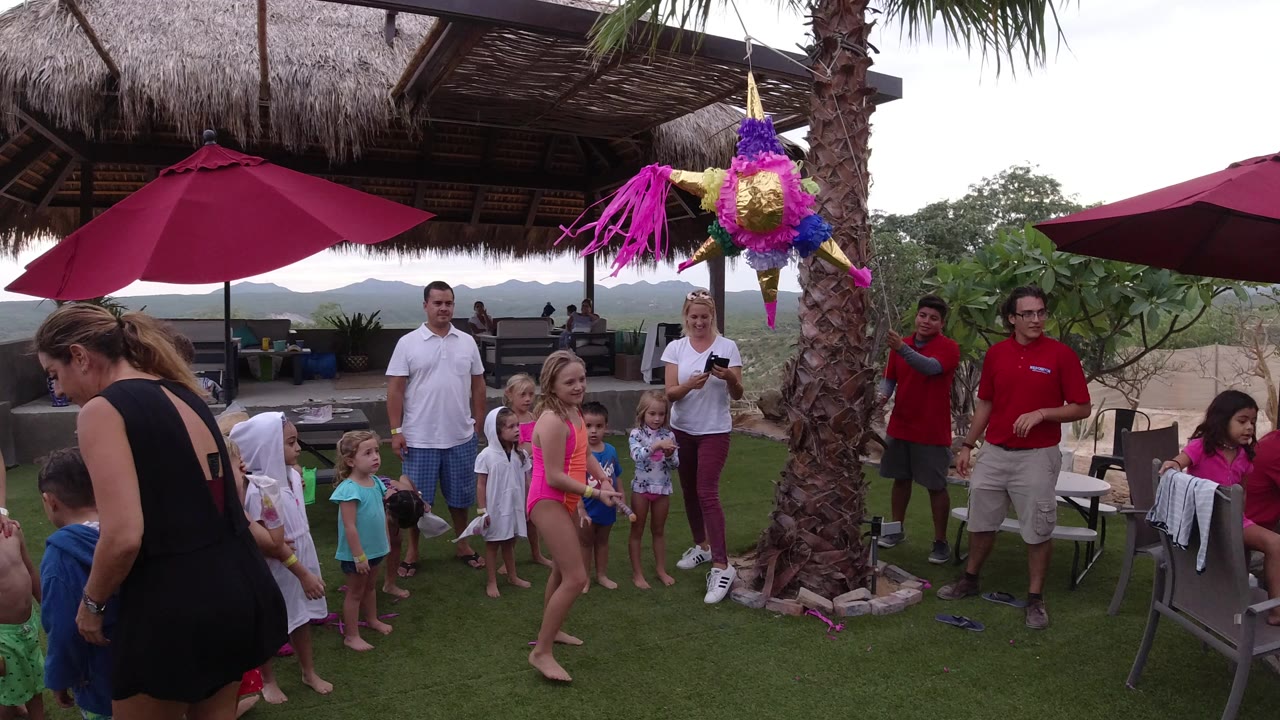 Children and adults gather around a hanging pinata on an outdoor patio.