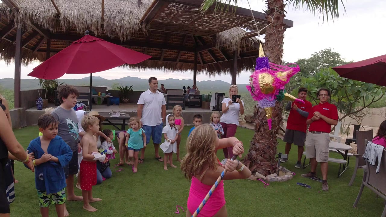 At a Los Cabos outdoor birthday party, a girl in a pink swimsuit faces a hanging pink and purple pinata while children and adults watch under red umbrellas.