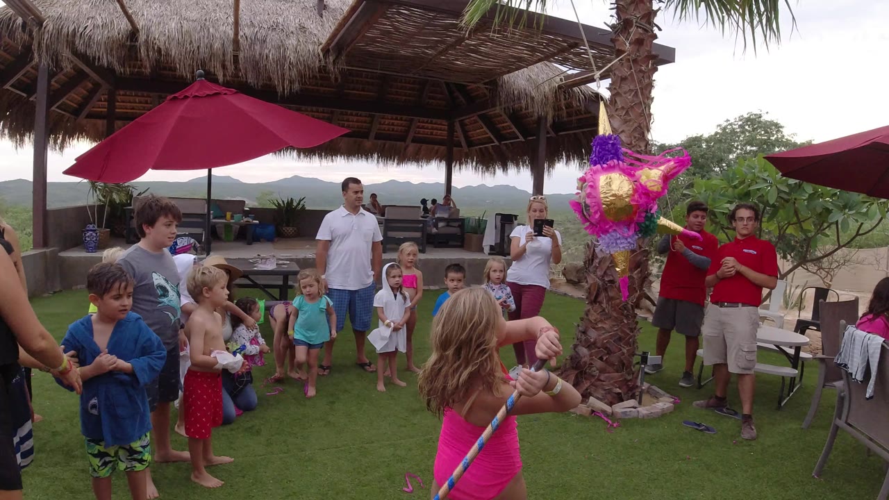 The girl holds the striped stick in the foreground as the colorful pinata hangs near a palm trunk, with adults and children watching from the lawn and patio in Los Cabos.