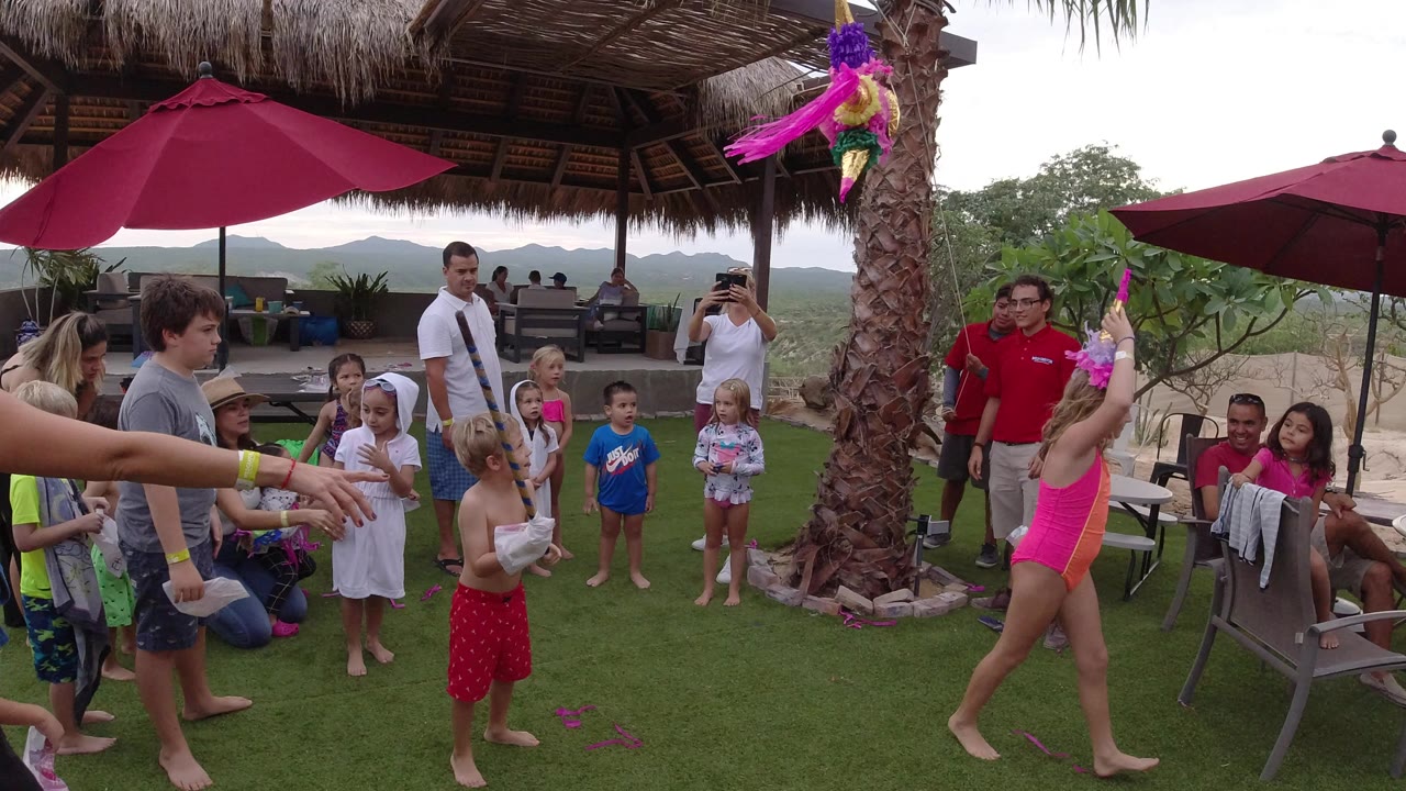 Child swings a stick at a hanging pinata while children and adults watch on a lawn.