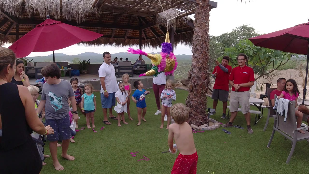 Children and adults gather under umbrellas near a hanging pinata.