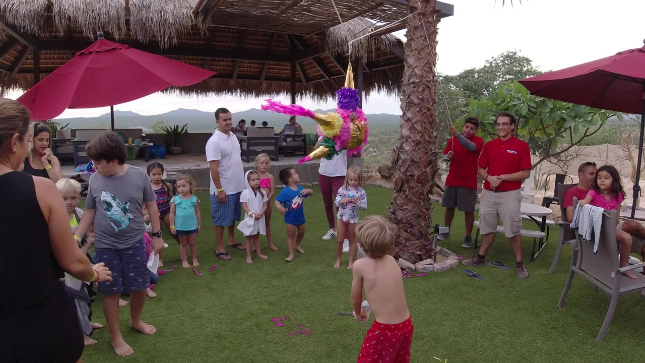 In Los Cabos, Mexico, a boy in a gray shirt stands near a line of children while adults watch beside the hanging pink and purple pinata.