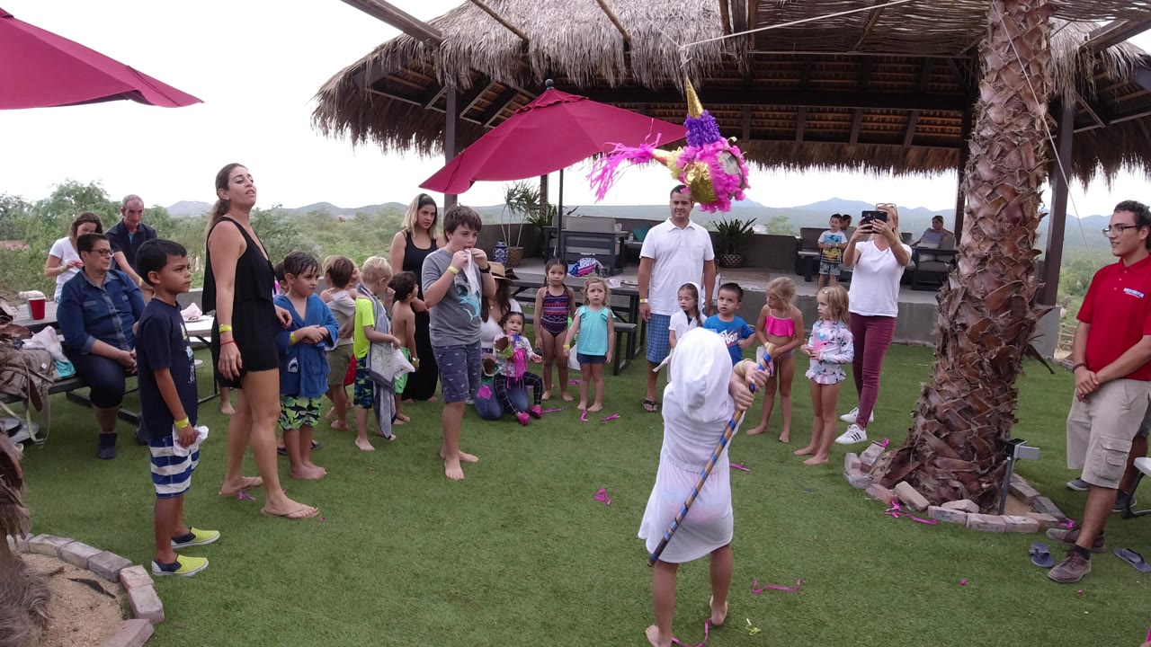 The child remains under the suspended pinata in Los Cabos while a semicircle of children and adults watches near red umbrellas and a thatched patio roof.