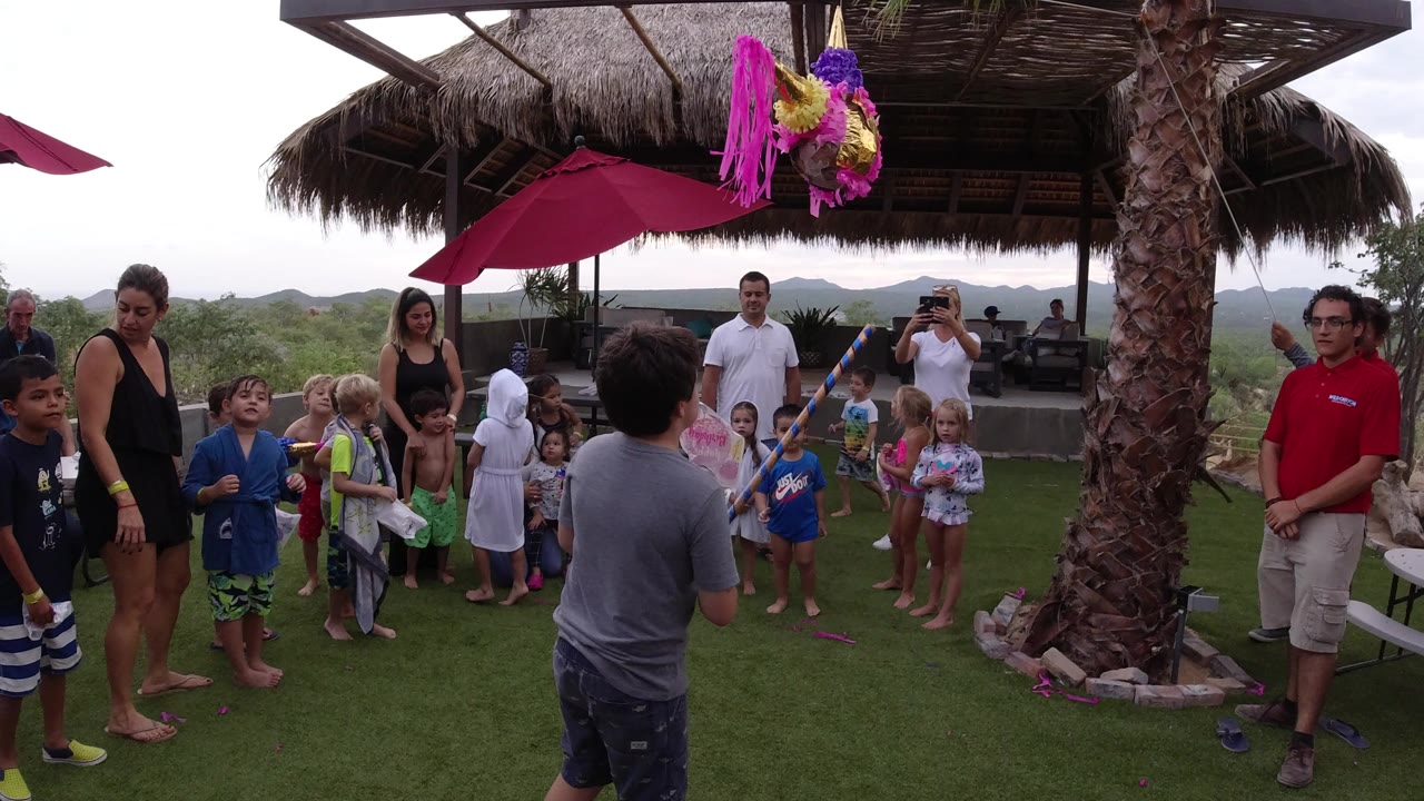 In Los Cabos, Mexico, a boy stands on grass facing a bright pink piñata while children and adults gather under a shaded outdoor patio.