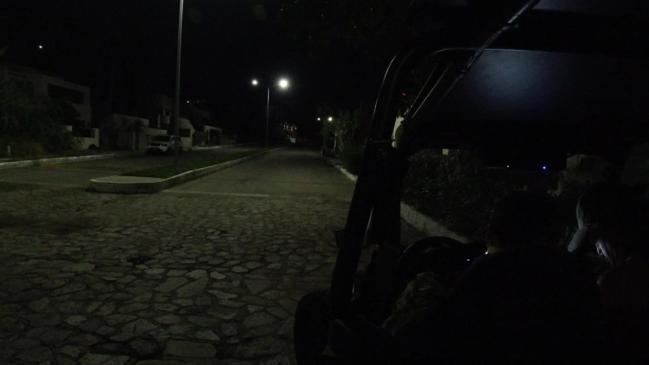 Opening frame of a dark Los Cabos cobblestone street seen from the rear of a motorized cart, with streetlights ahead, parked vehicles at left, and no clearly visible people.
