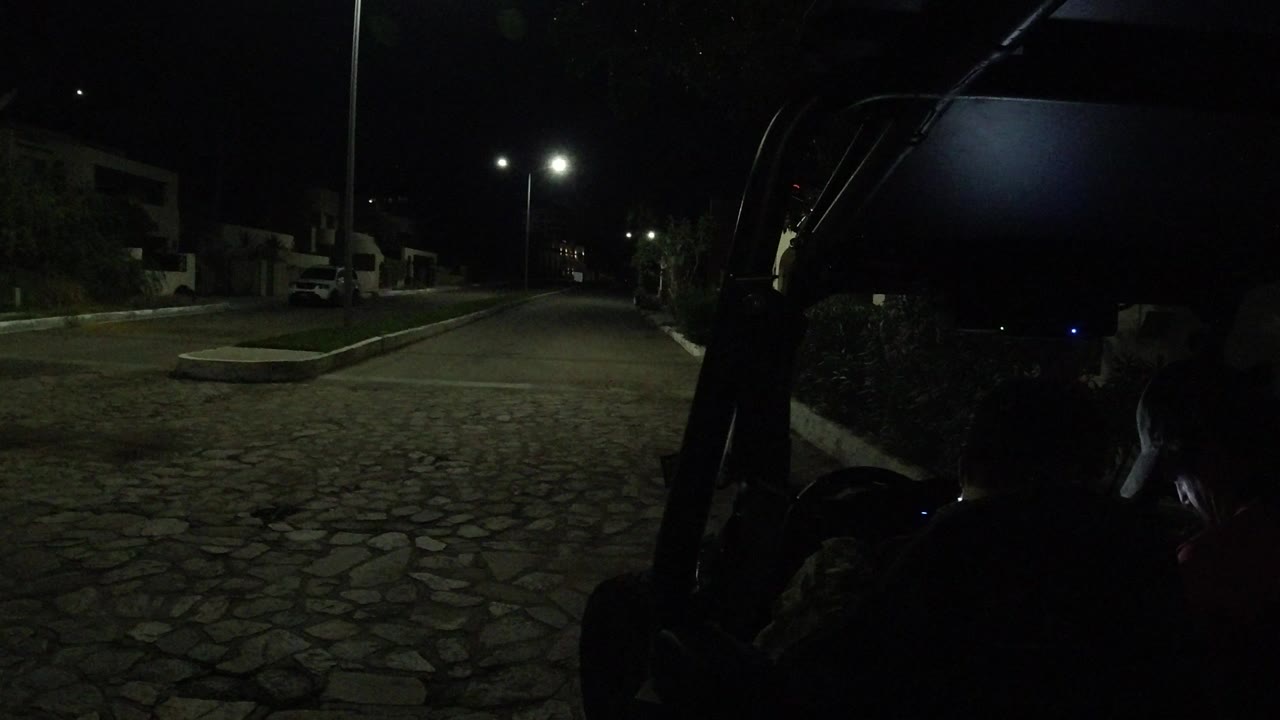 Rear-mounted cart view in Los Cabos with the cobblestone road centered, twin streetlights bright in the distance, and the cart roof edge dark across the foreground.