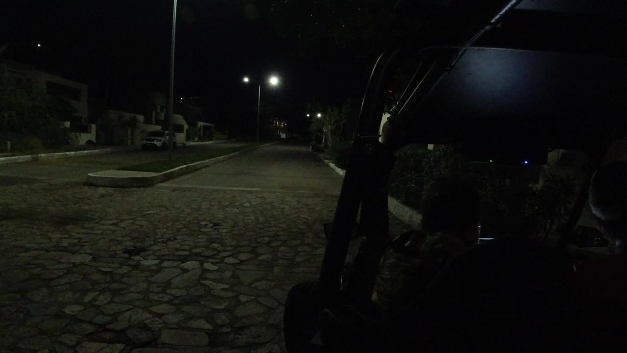 Los Cabos night frame with the Wrangler forming a dark foreground edge, a mostly empty stone road ahead, and streetlights illuminating parked vehicles and buildings.