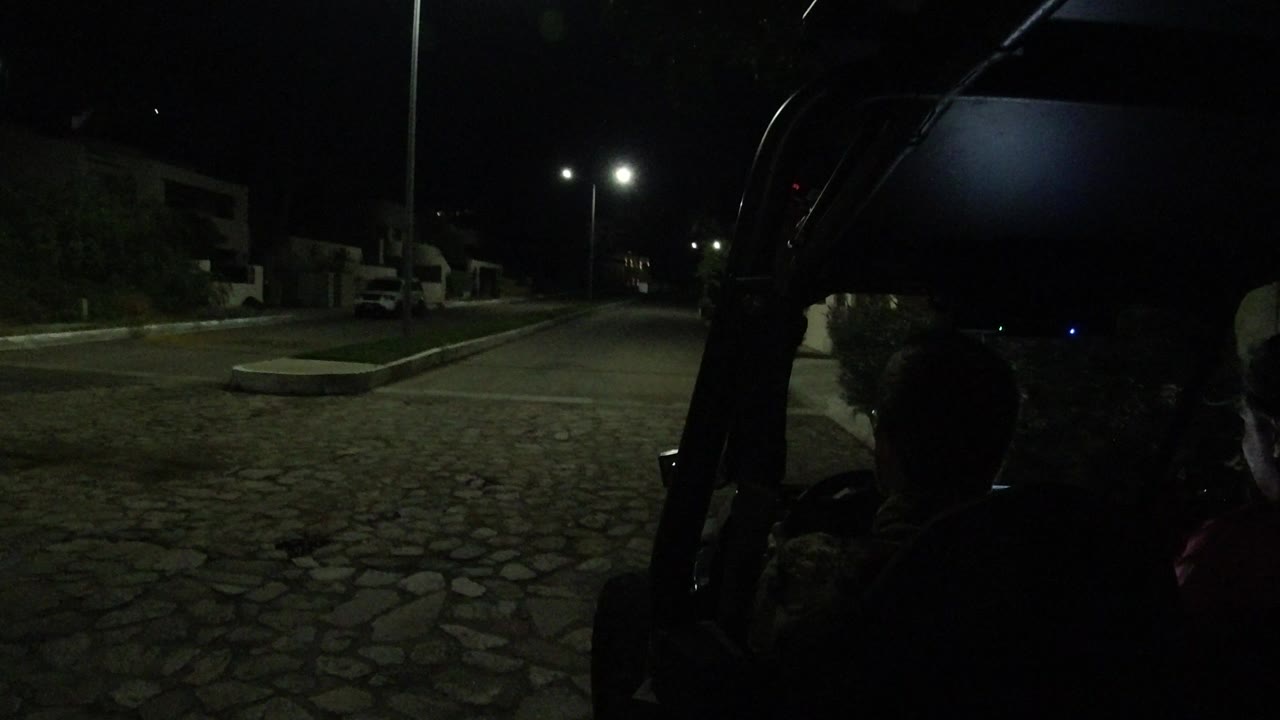 A nighttime Los Cabos road scene shows the open vehicle frame in the foreground, two riders in silhouette, a divided curb area, and streetlights ahead.