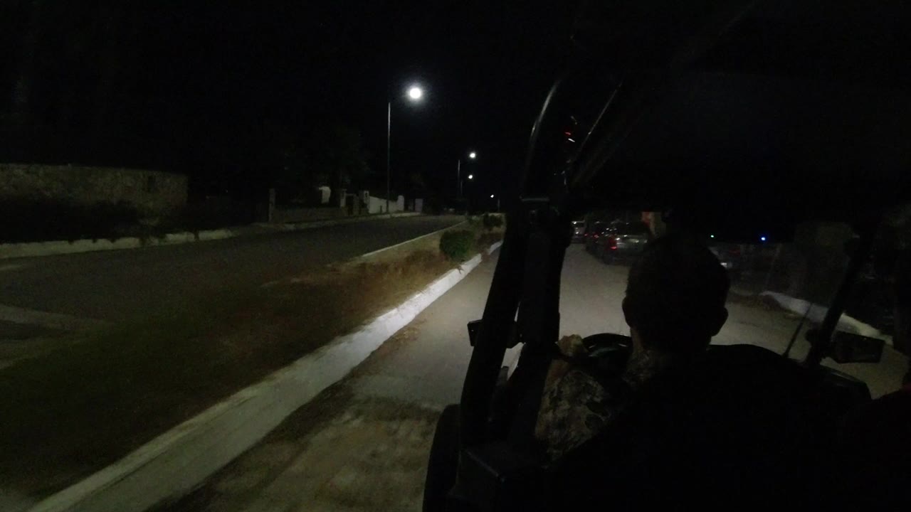 A dark Los Cabos road is seen from behind a silhouetted rider, with a curb, shrubs, parked cars, and streetlights ahead.