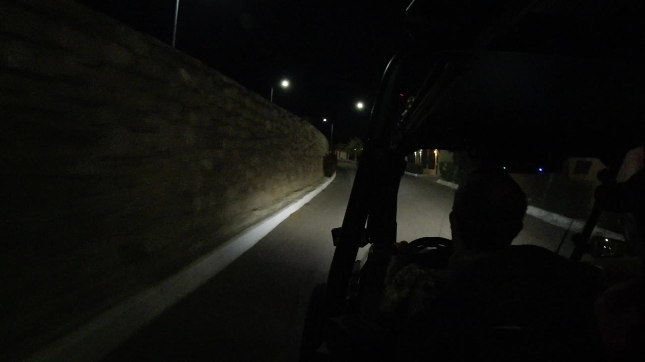 A nighttime Los Cabos road continues along the stone wall, with a bright streetlamp in the distance and the vehicle structure silhouetted at left.