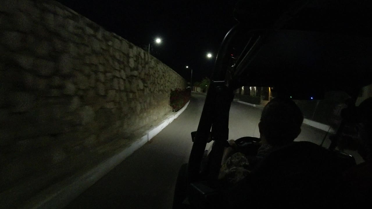 The road in Los Cabos passes close to the rough stone wall, with the seated silhouette centered low in frame and streetlights marking the route ahead.