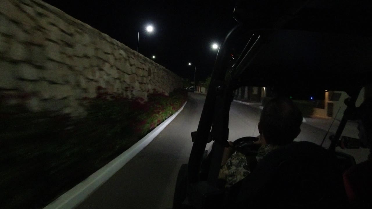 A dark Los Cabos road runs past the stone wall with bright overhead lamps, a parked vehicle ahead, and a shadowed occupant visible from behind.