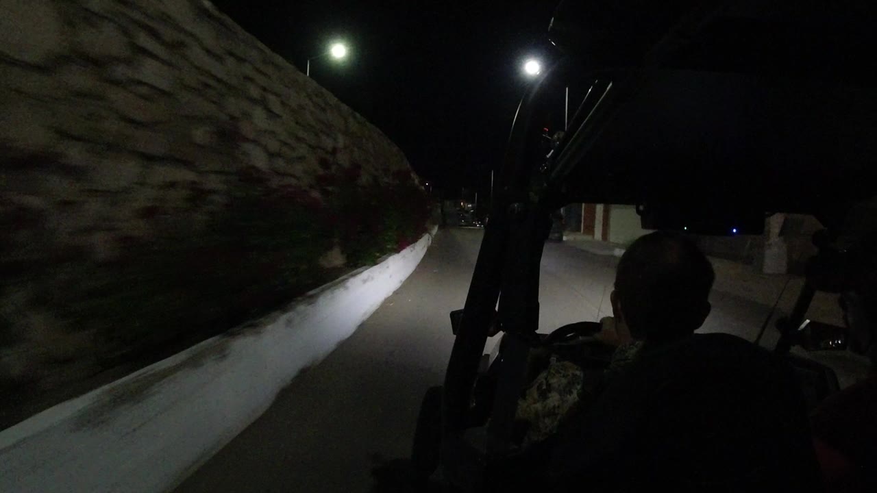 Inside a Jeep at night in Los Cabos, a dark driver silhouette sits ahead while headlights light a curb and stone wall along the road.