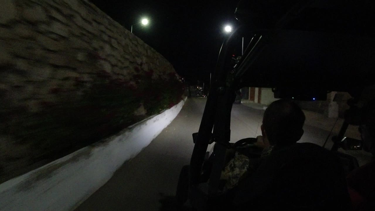The Los Cabos road continues past a rough stone wall, with the Jeep frame and driver silhouette in the dark foreground and streetlights ahead.