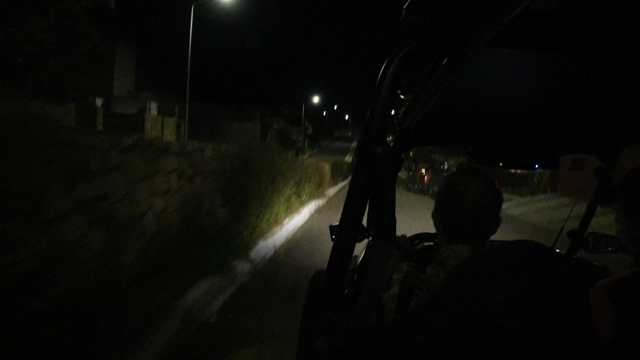 The vehicle continues along a narrow Los Cabos night road, with the headlight beam catching the wall and brush while passenger silhouettes remain in the foreground.