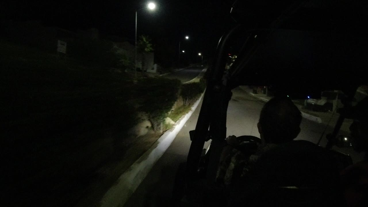 A Los Cabos night street is viewed from the golf cart, with a passenger silhouette centered and streetlights reflecting on the curb and wall ahead.