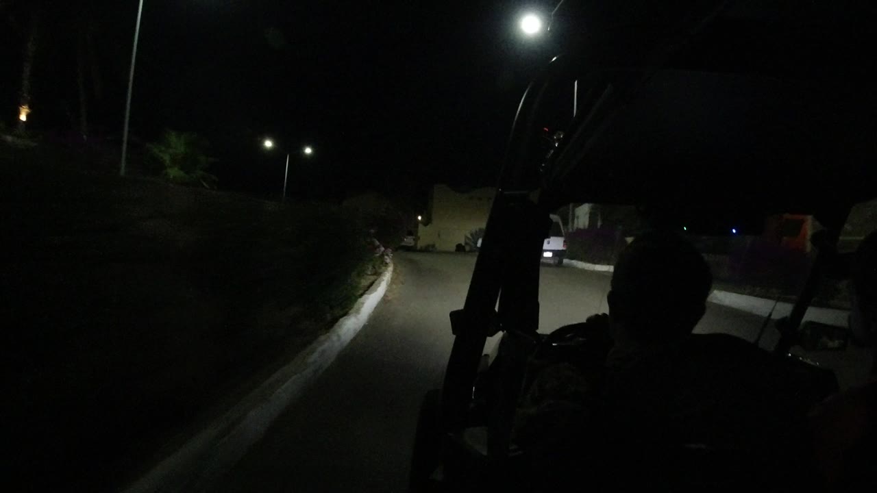 From behind the silhouetted driver, the Los Cabos road curves past hedges and parked vehicles lit by bright streetlights in the night.