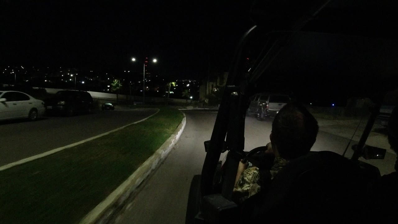 The driver remains in shadow at the right edge while the Los Cabos roadway curves ahead beside a lit grass island and parked vehicles.