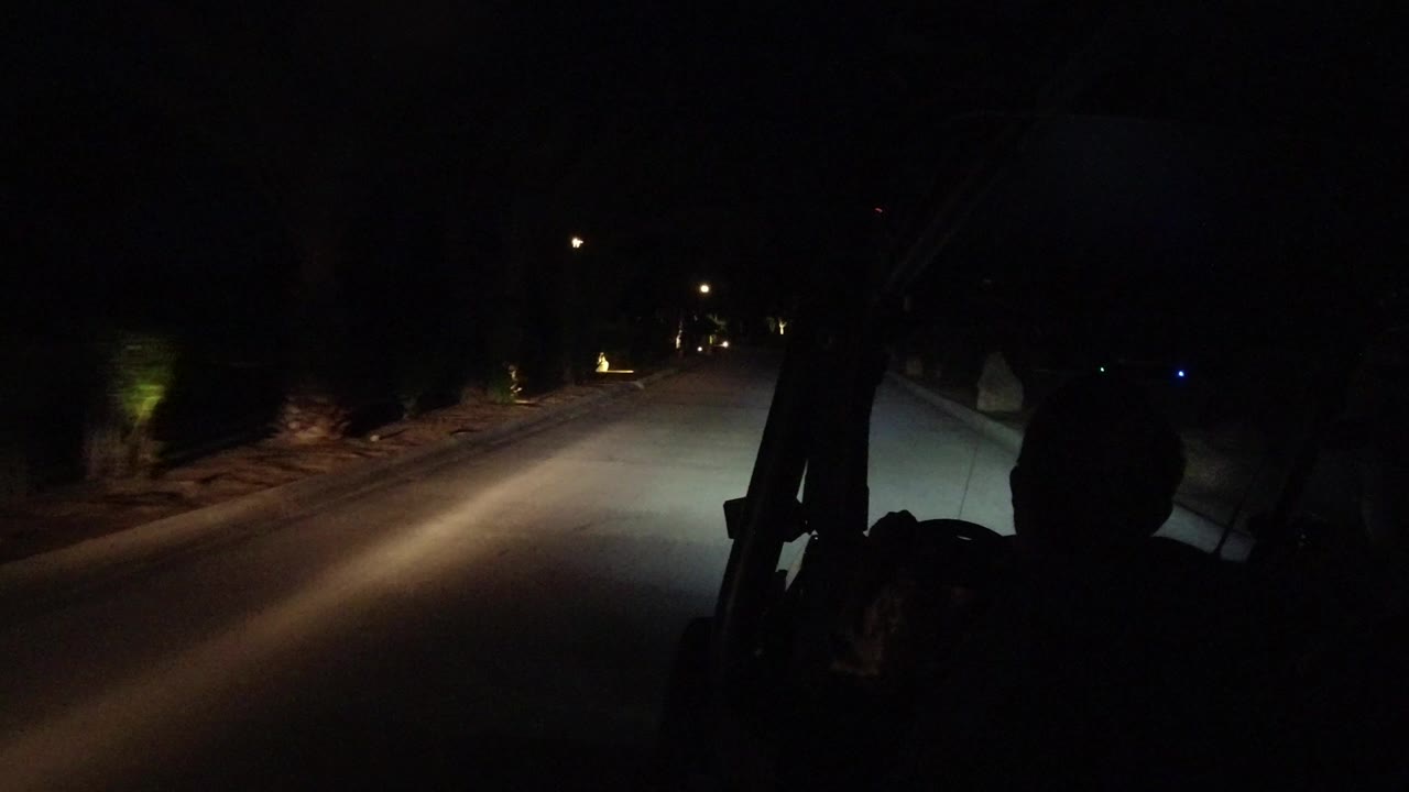 Night road in Los Cabos, Mexico seen from inside or behind a Wrangler, with headlight beams on pavement, roadside planting, and no clear faces visible.