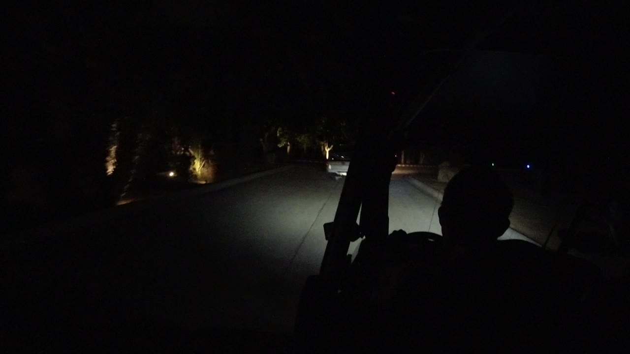 A dim Los Cabos road curves through darkness, with headlights lighting the pavement and small landscape lamps marking trees along the roadside, with no clear visible people.