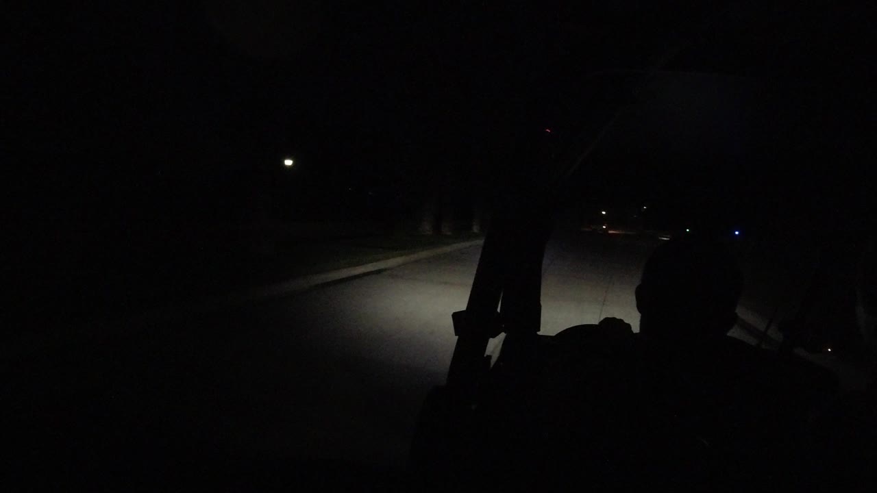 A near-black Los Cabos road view shows the headlights reaching farther down the lane, with a small overhead light and dark foreground framing still obscuring details.