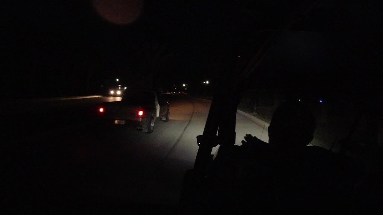 Los Cabos night driving frame with the road lit by headlights, a car ahead showing red taillights, and a dark passenger silhouette in the foreground.