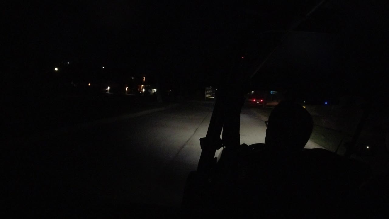 A dark vehicle interior in Los Cabos, Mexico looks toward a dim street, with silhouetted passengers and headlights spreading across the pavement.