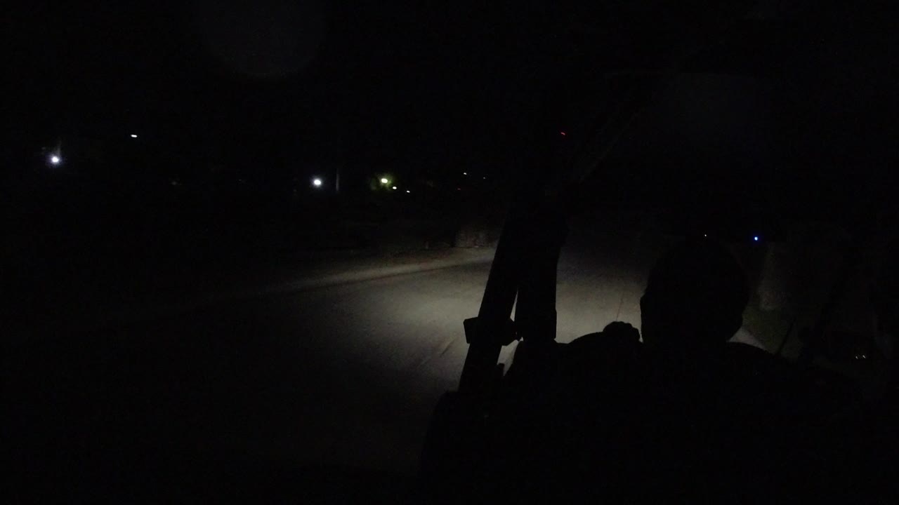 A very dark Los Cabos road is seen from inside or on a vehicle, with headlights lighting the pavement and passenger silhouettes in the foreground.