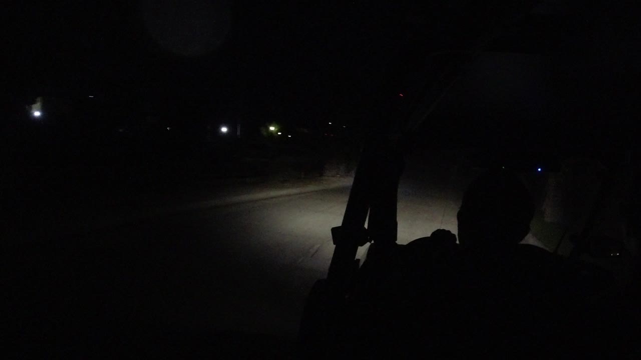 The vehicle continues along a nearly empty Los Cabos road, with a bright headlight patch on the pavement and dark rider silhouettes near the frame edge.