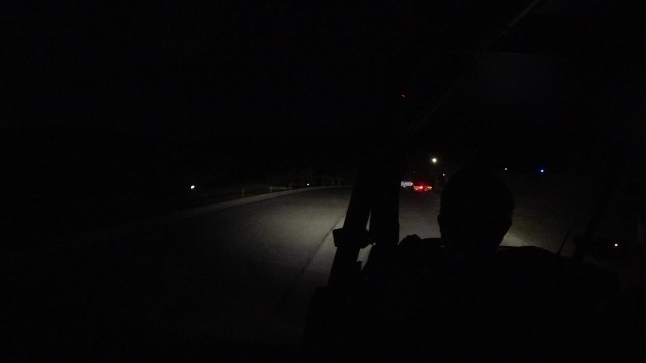 At 0:01, the headlights brighten the right side of the road while the foreground passenger remains a dark silhouette against sparse roadside lights in Los Cabos.