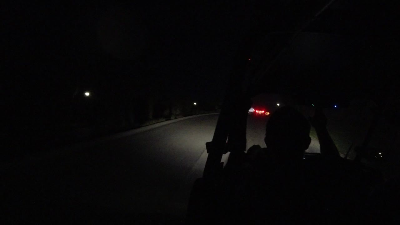 A dark interior view in Los Cabos, Mexico shows a silhouetted driver facing a night road lit by headlights, with red taillights visible ahead.