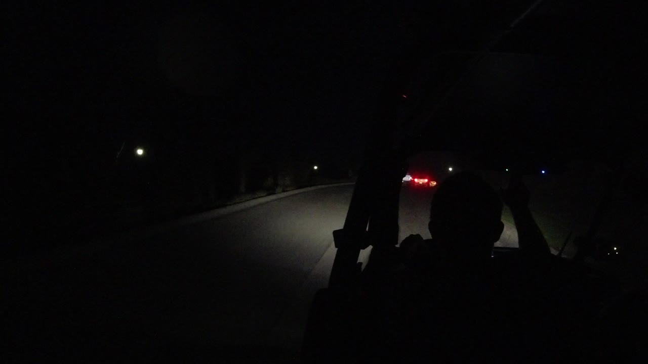 A silhouetted driver remains centered in the foreground while headlights reveal a curving dark road in Los Cabos, Mexico and small lights appear in the distance.