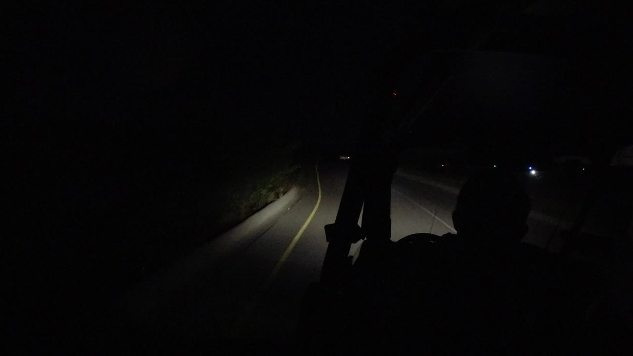 The Los Cabos night road curves through the headlight beam while the driver remains a dark seated silhouette in the foreground.