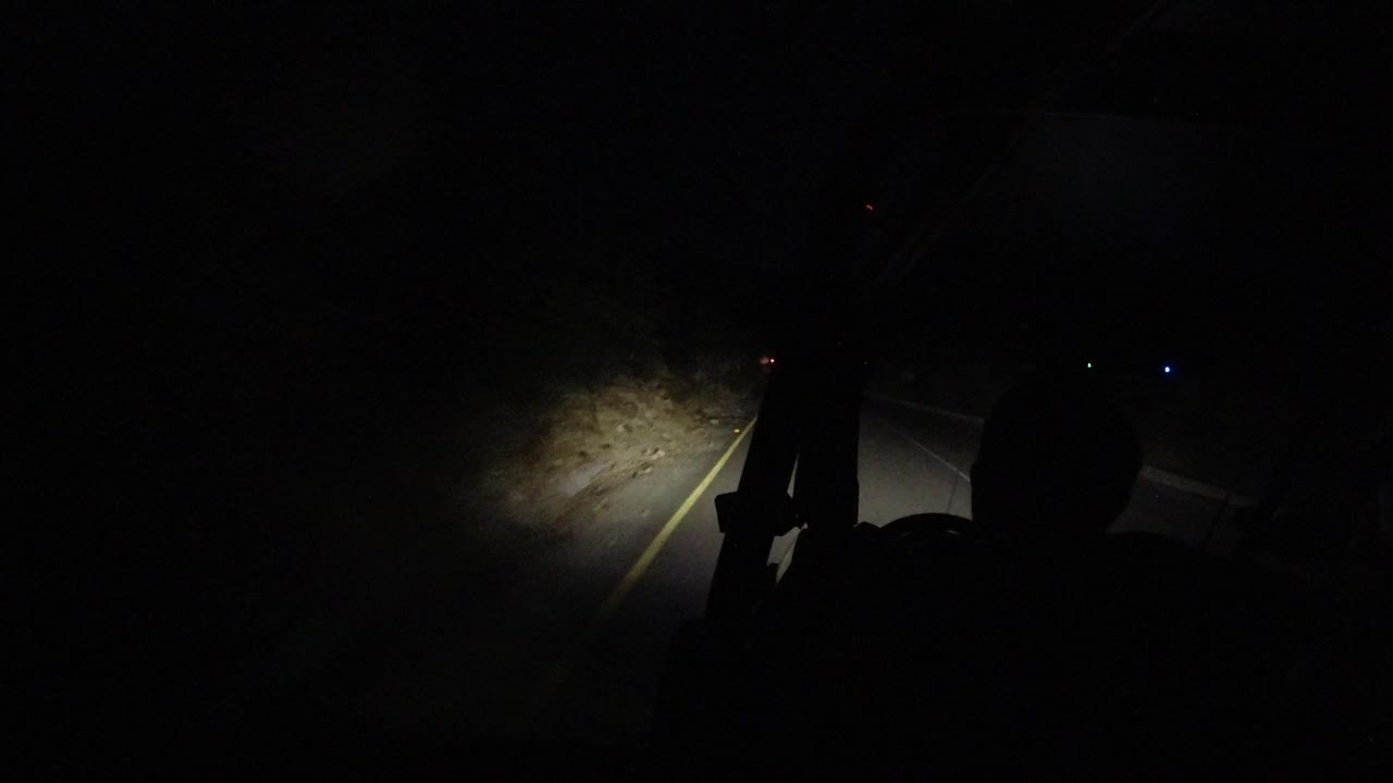 The Los Cabos night road remains mostly black while the light falls farther along the roadside dirt and yellow line, with the jeep frame silhouetted and no visible people.