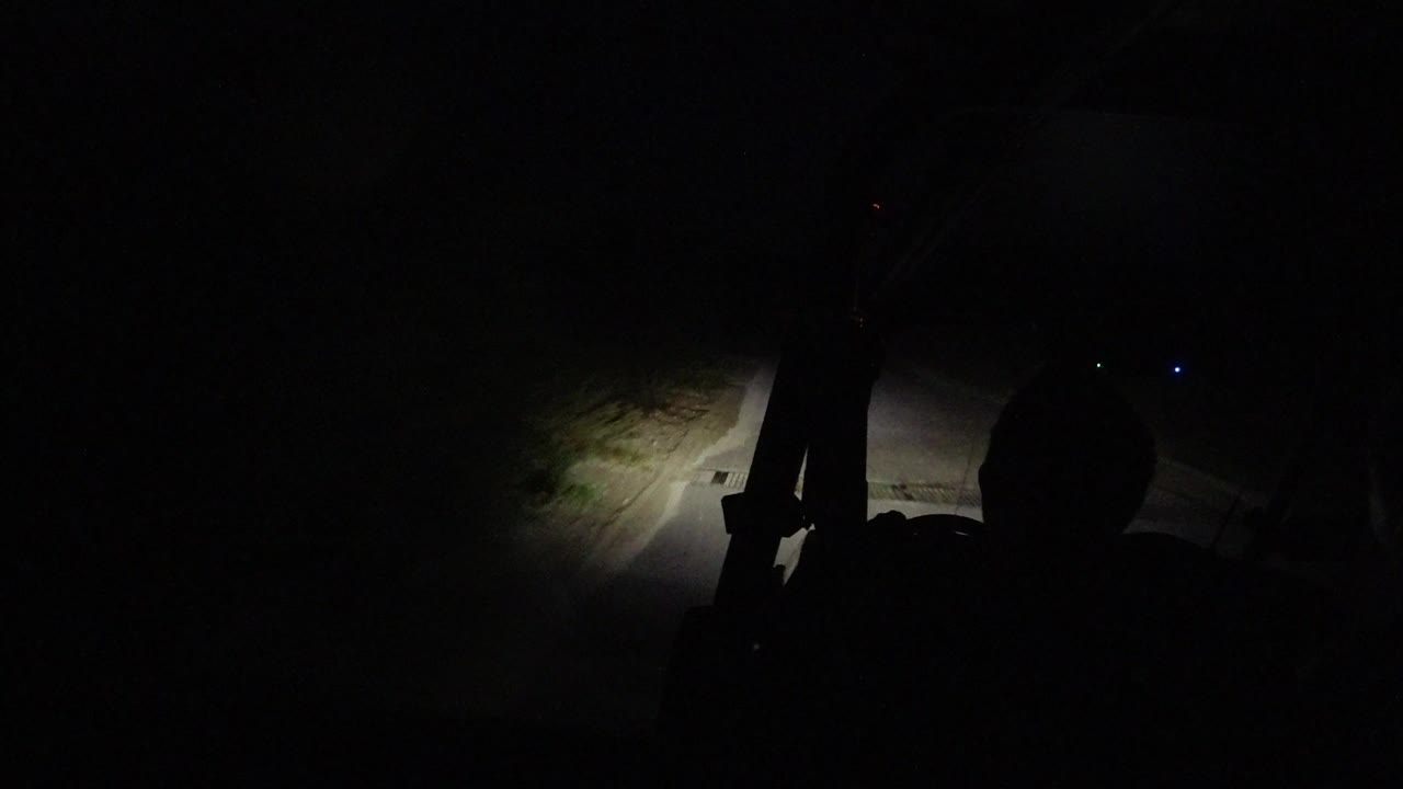 At 0:00 in Los Cabos, Mexico, a dark vehicle foreground frames a narrow road and sandy shoulder lit by headlights, with no clearly visible people.