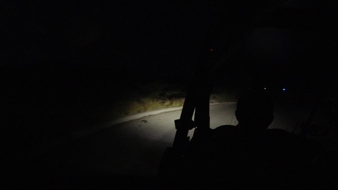 Inside the vehicle, the headlight beam reaches a curving empty road in Los Cabos, Mexico while the front-seat figure remains mostly in silhouette.