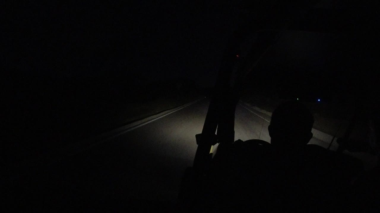 At 0:01 in Los Cabos, Mexico, the road remains mostly black while the headlights catch a pale lane edge beside a seated human silhouette.