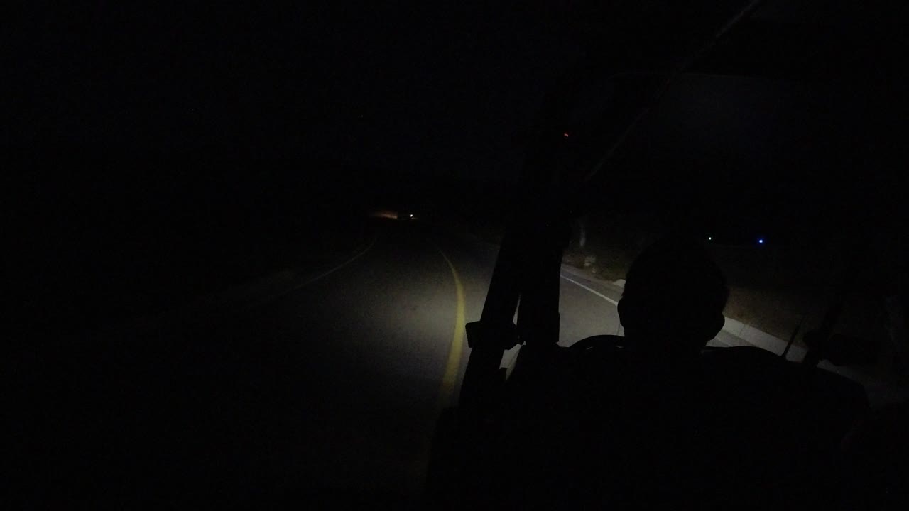A low-light Los Cabos highway view shows a curving lane line and a small patch of illuminated shoulder, with no visible people.