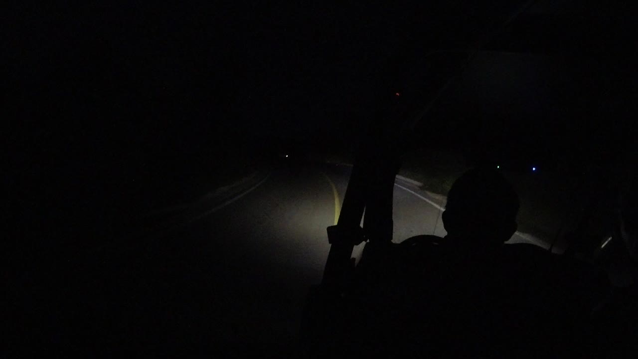 A dark nighttime view in Los Cabos, Mexico shows an ATV rider silhouette in the foreground and yellow road stripes lit by headlights ahead.