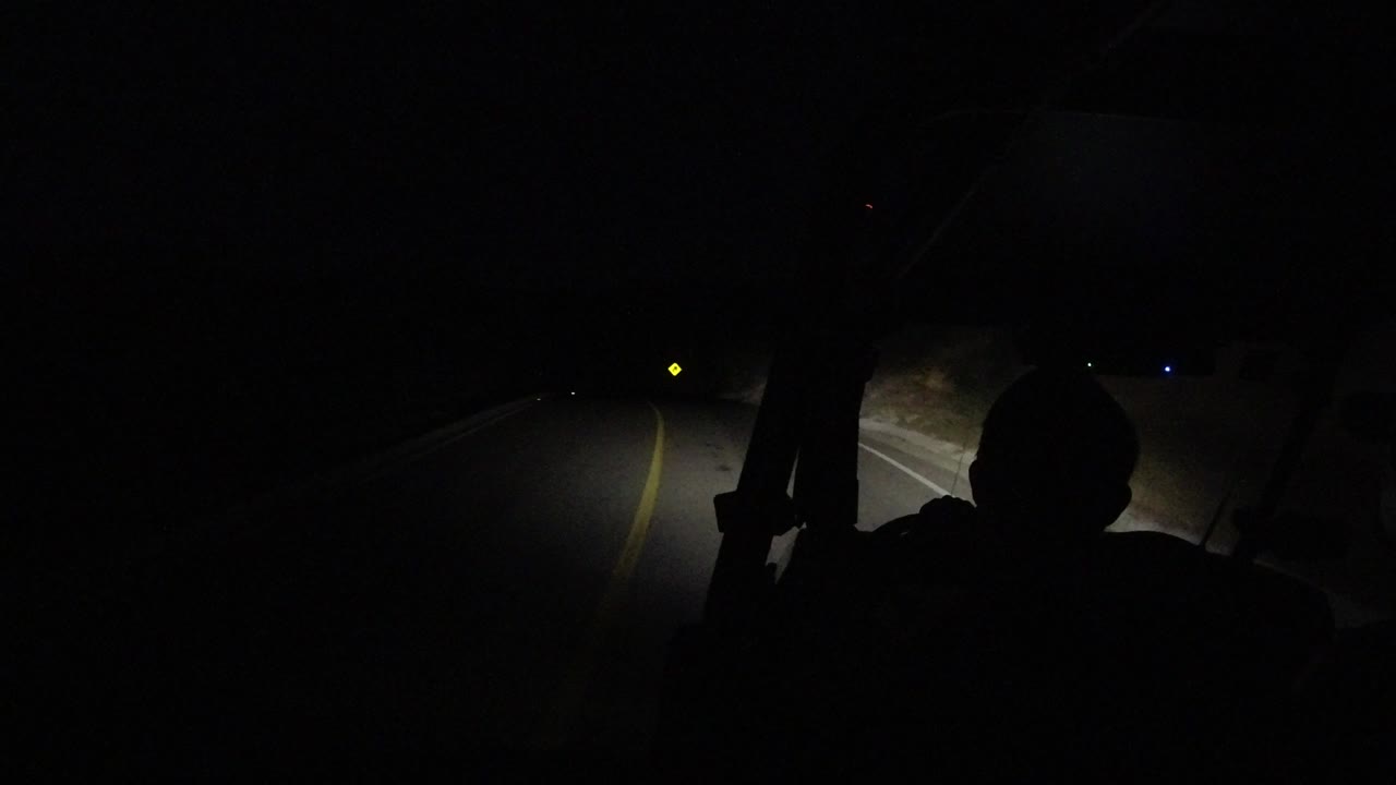 At 0:01 in Los Cabos, Mexico, the dark occupant silhouette remains centered while the headlight beam catches the yellow road line and pale shoulder.