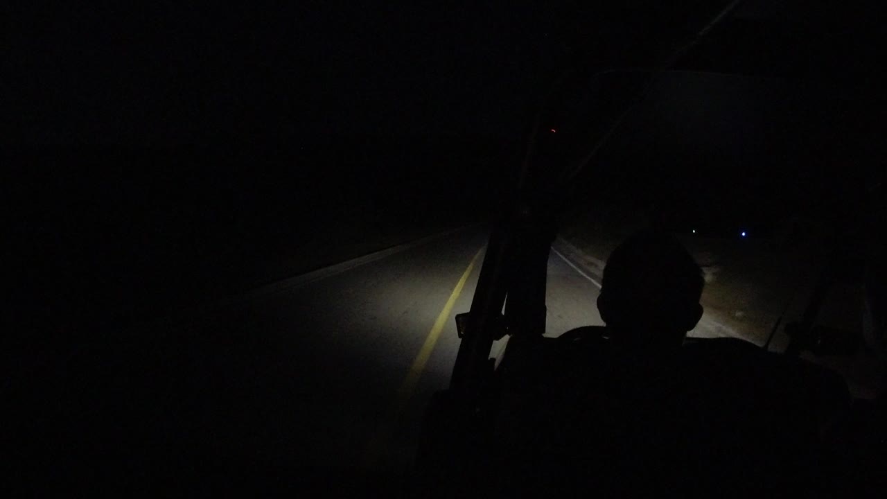 Start of the Los Cabos clip with headlights lighting a narrow stretch of unlighted road through a dark vehicle frame, no clearly visible people.