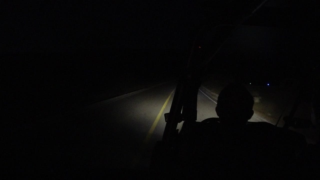 A forward view through the dark vehicle interior shows the Los Cabos road lane and shoulder caught in the headlights, with no clearly visible people.
