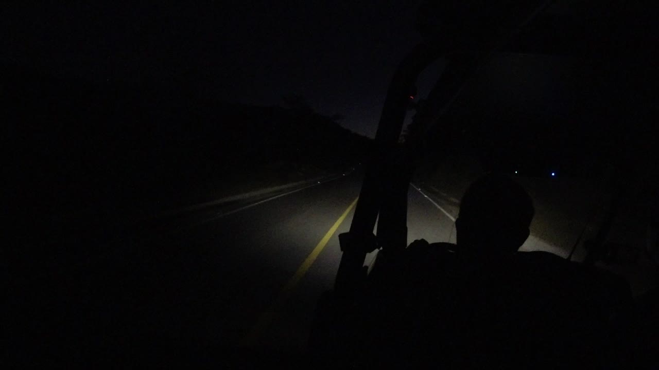 At 0:00 in Los Cabos, Mexico, headlights pick out a yellow road line while a dark driver-like silhouette sits in the foreground with no recognizable face.