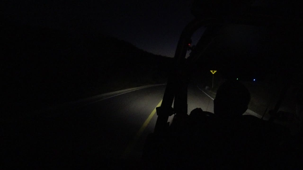 A dark Los Cabos road at night is viewed from a vehicle, with headlights catching the yellow edge line, hillside silhouettes, and a dark occupant silhouette without a visible face.