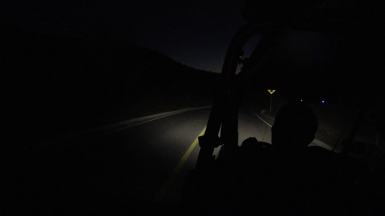 At 0:01, the Los Cabos night road remains mostly black, with the headlight beam stronger on the pavement and a rounded occupant silhouette visible in the lower right.