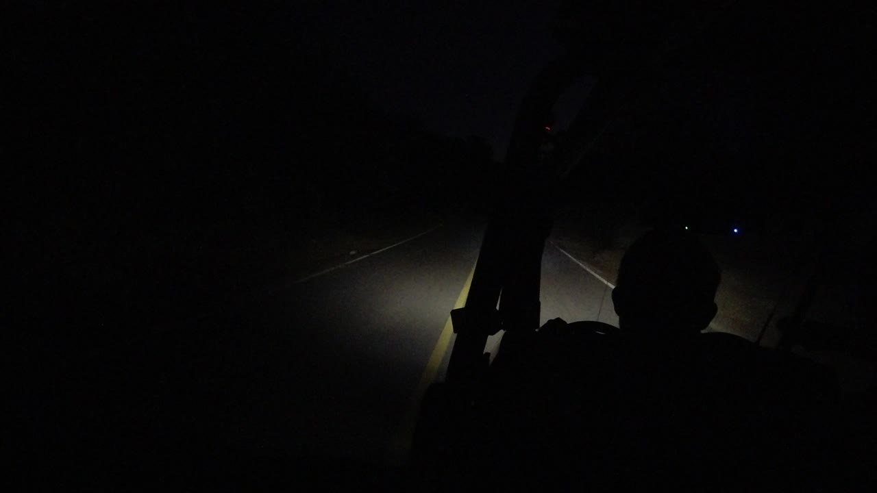 A headlight pool falls across the road in Los Cabos as the passenger silhouette remains near the bottom right and small lights appear in the distance.