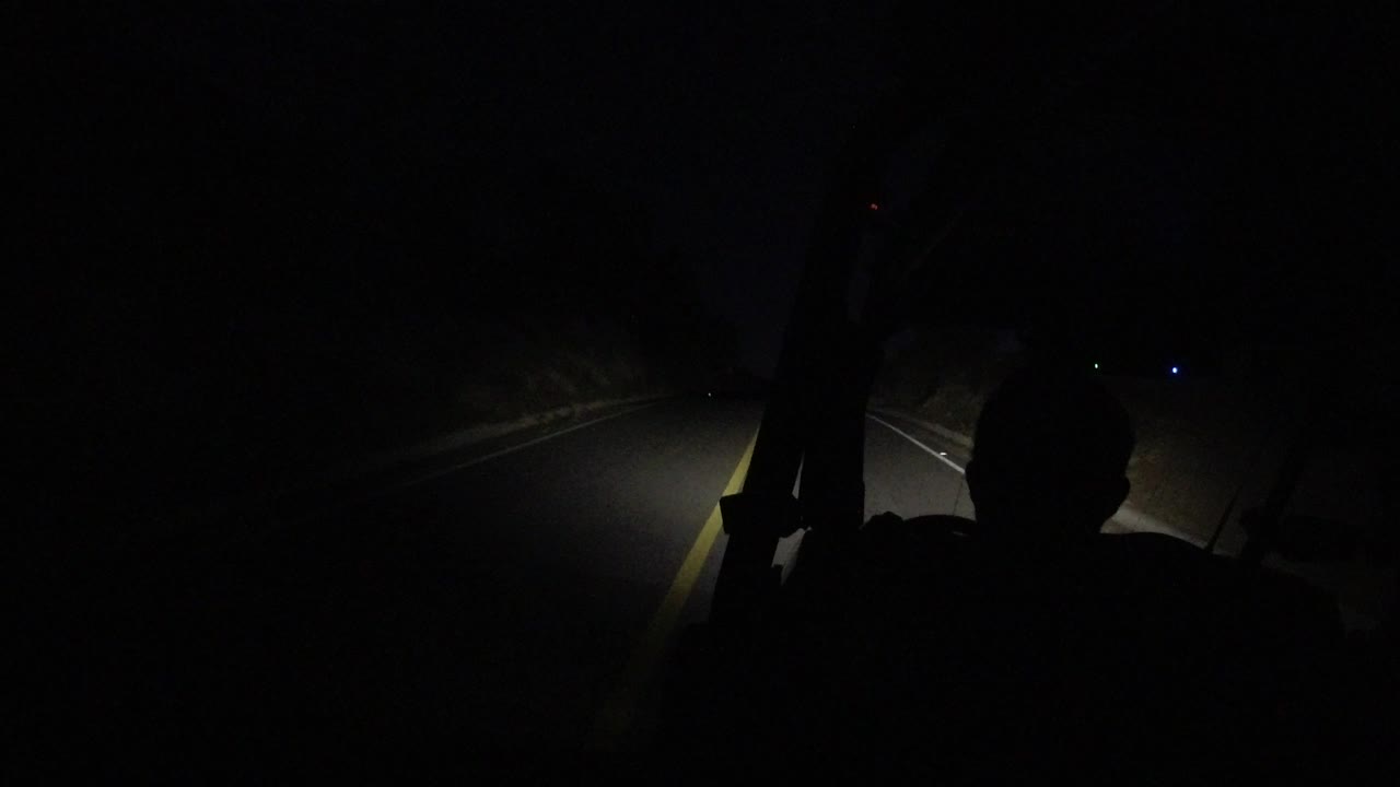 The Los Cabos night drive continues with the driver silhouette centered lower in frame and the headlights catching the lane line and roadside shoulder.