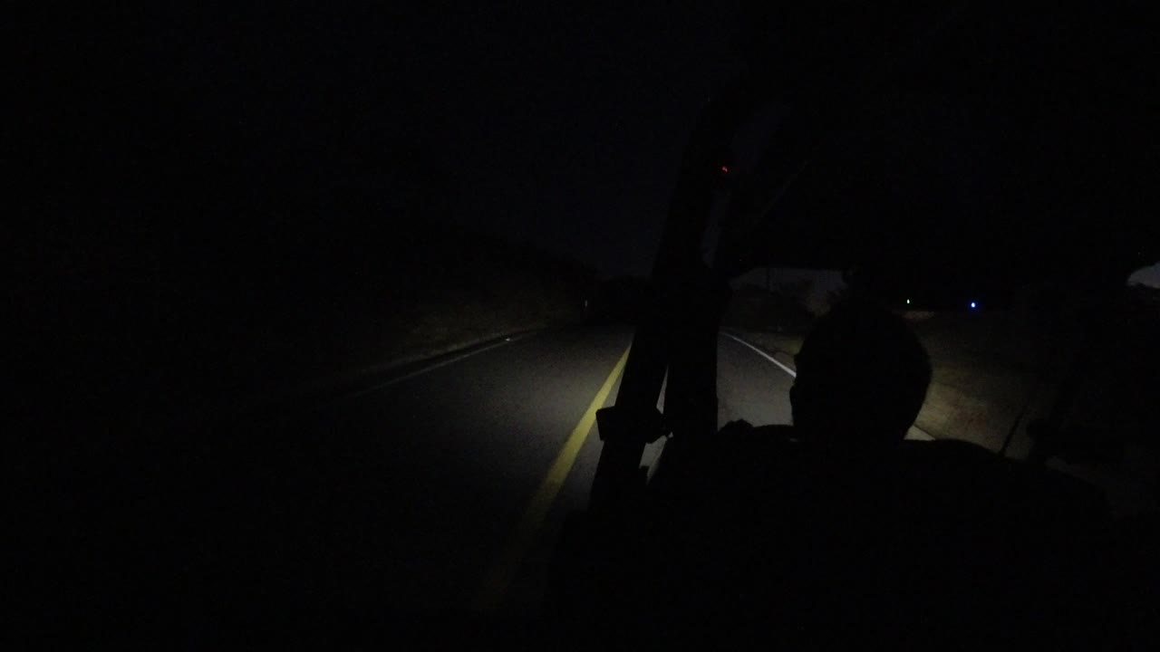 A forward road view in Los Cabos, Mexico shows headlights spreading across pavement and a roadside edge, with no clearly visible people.